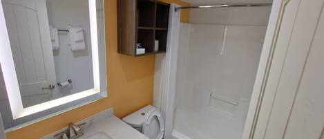 Un baño con lavamanos, inodoro y bañera blancos. Hay un espejo sobre el lavamanos y un estante sobre el inodoro.