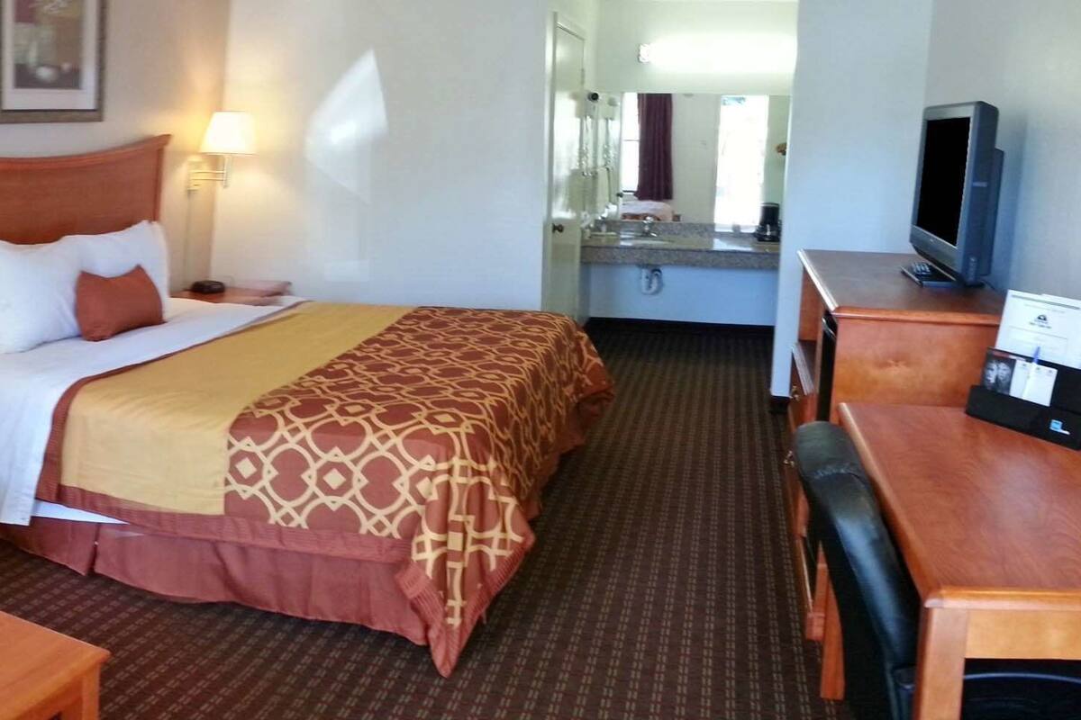 A hotel room with a bed, a desk, a chair, a TV, and a bathroom visible in the background.