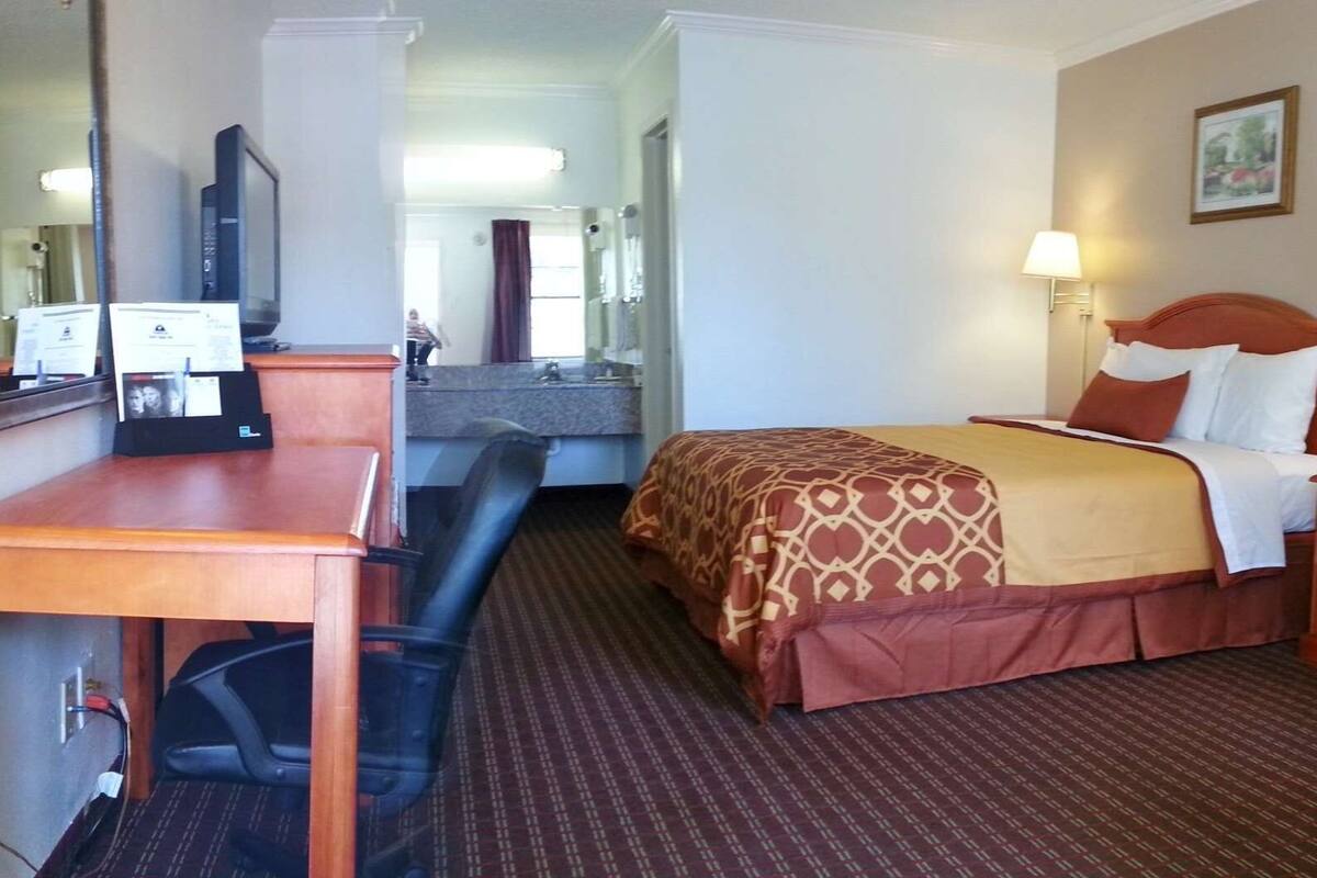 A hotel room with a bed, desk, chair, and a small table with a lamp.