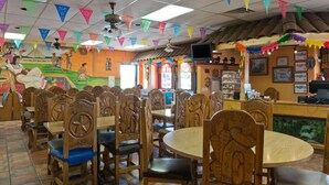 Restaurante com cadeiras e mesas de madeira, decorações coloridas e um mural na parede.