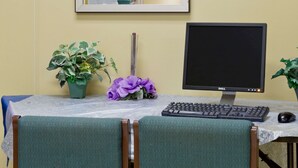Uma escrivaninha com um monitor, teclado e rato. Há duas cadeiras, um vaso de planta e um vaso com flores roxas.