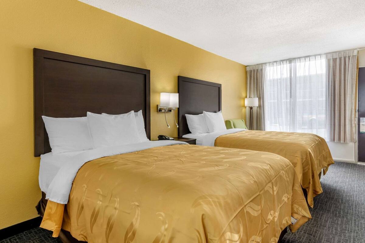 A hotel room with two beds, a nightstand, a lamp, and a window with curtains.