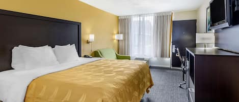 A hotel room with a large bed, a desk with a flat-screen TV, a chair, and a desk lamp.