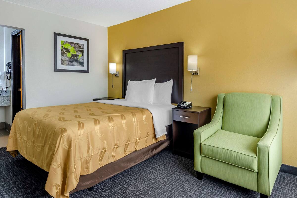 A hotel room with a bed, a green armchair, a nightstand with a phone, and a framed picture on the wall.