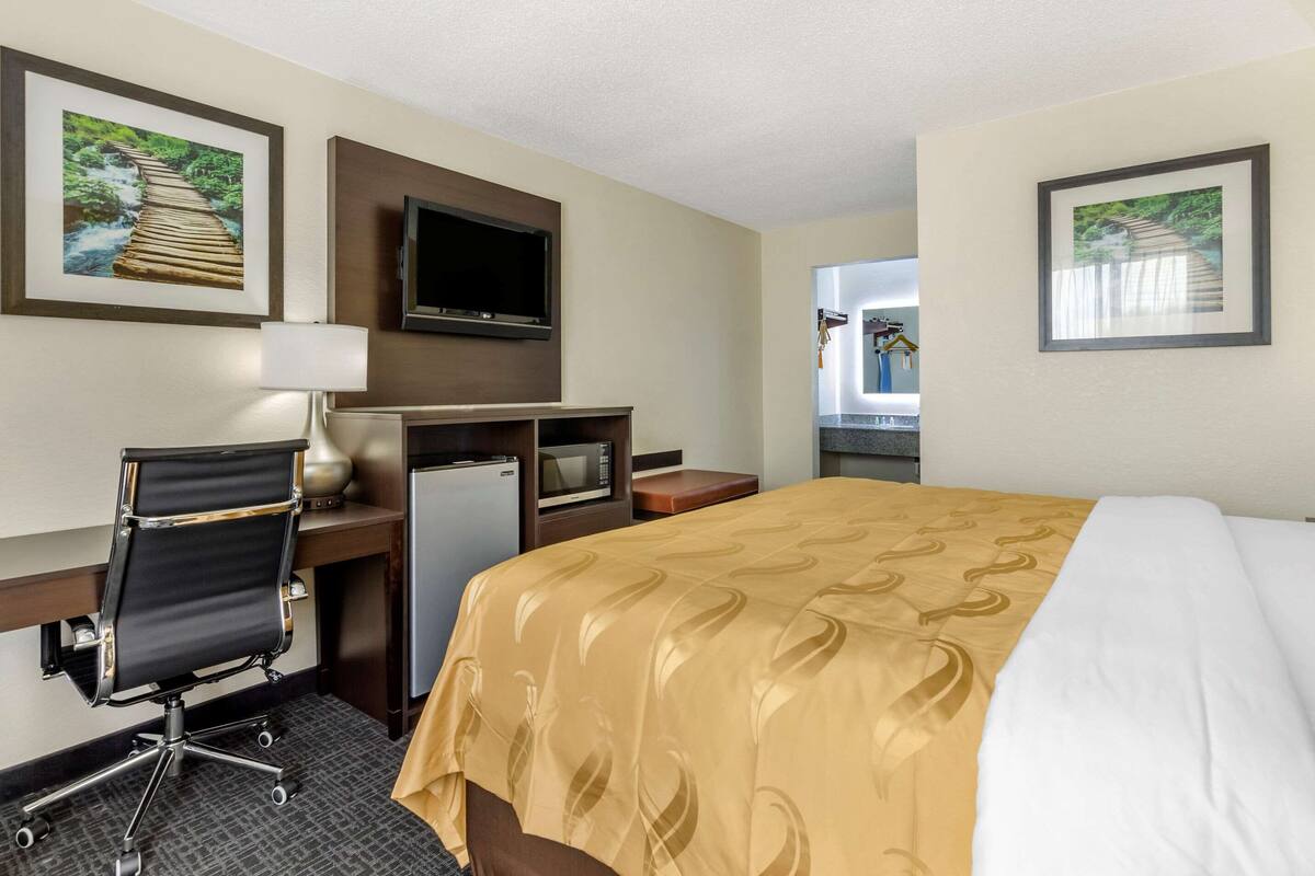 A hotel room with a bed, desk, chair, TV, and a small kitchenette.