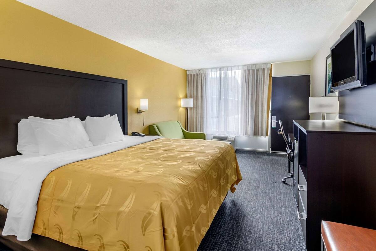 A hotel room with a bed, a desk, a chair, a television, and a desk lamp.