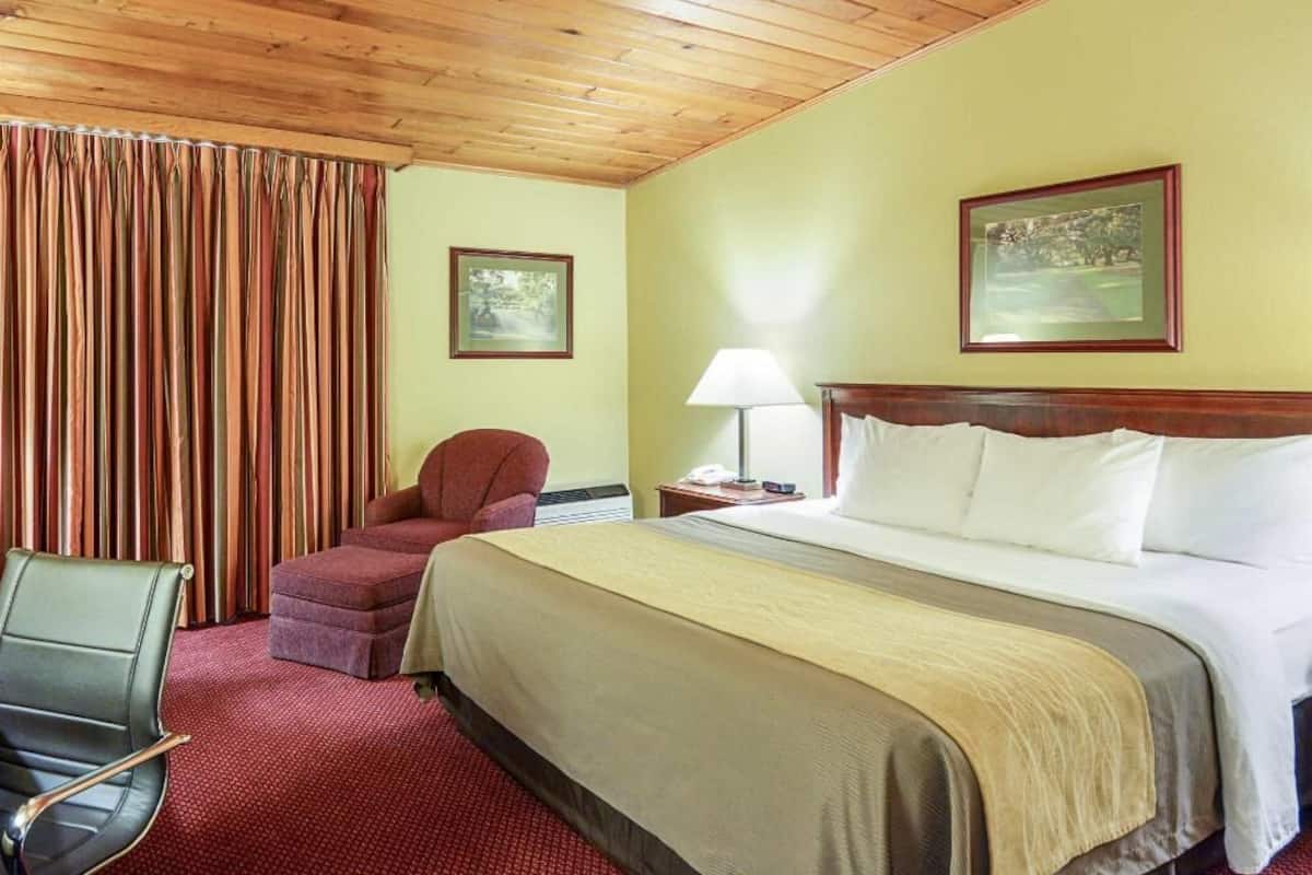 A hotel room with a wooden ceiling, a bed with white linens, a red chair, a desk with a lamp, and two framed pictures on the wall.