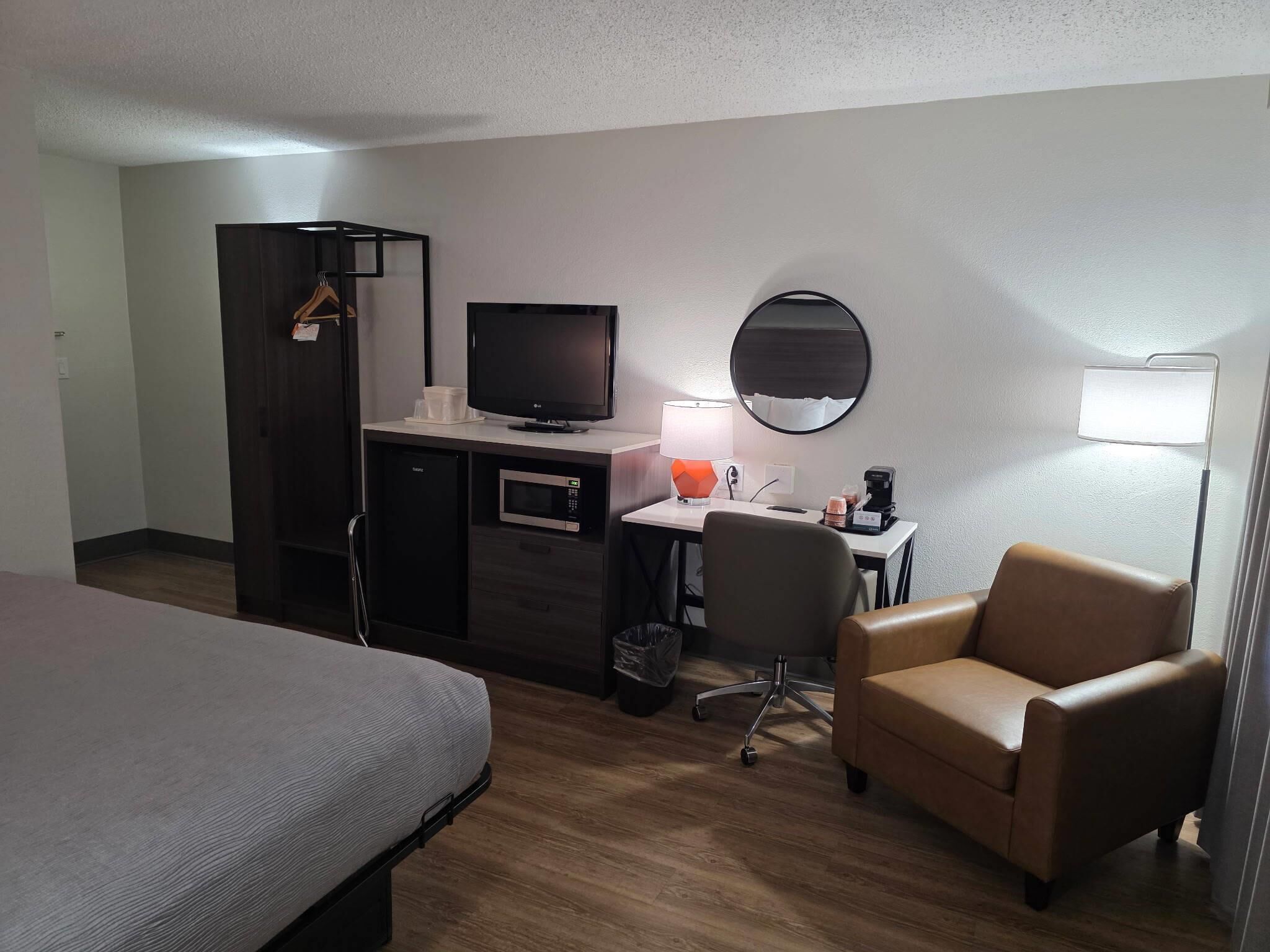 A hotel room with a bed, a desk with a computer, a chair, a TV, a lamp, and a coffee maker.