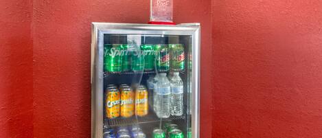 Máquina expendedora con diferentes bebidas, incluyendo Sprite, Crush Naranja y Pepsi, frente a una pared roja.