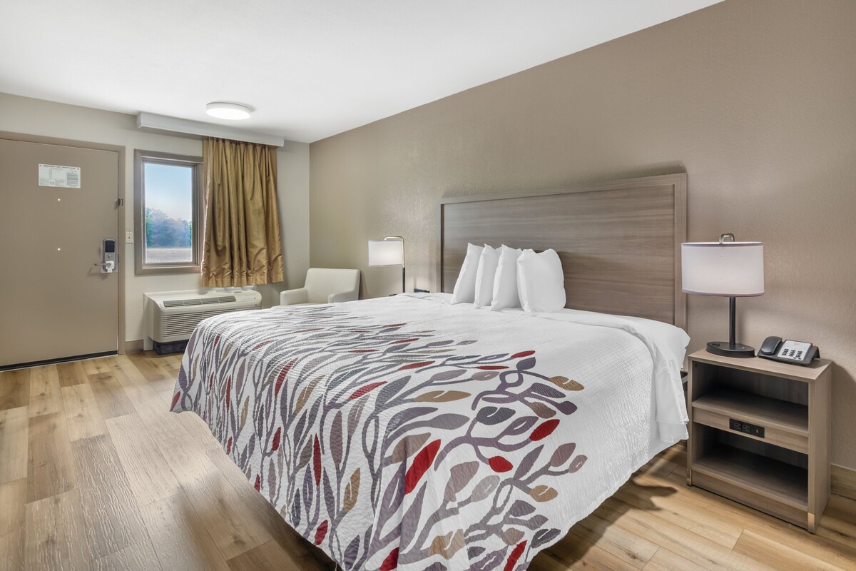 A hotel room with a large bed, two bedside tables with lamps, a window with curtains, and a door with a control panel.