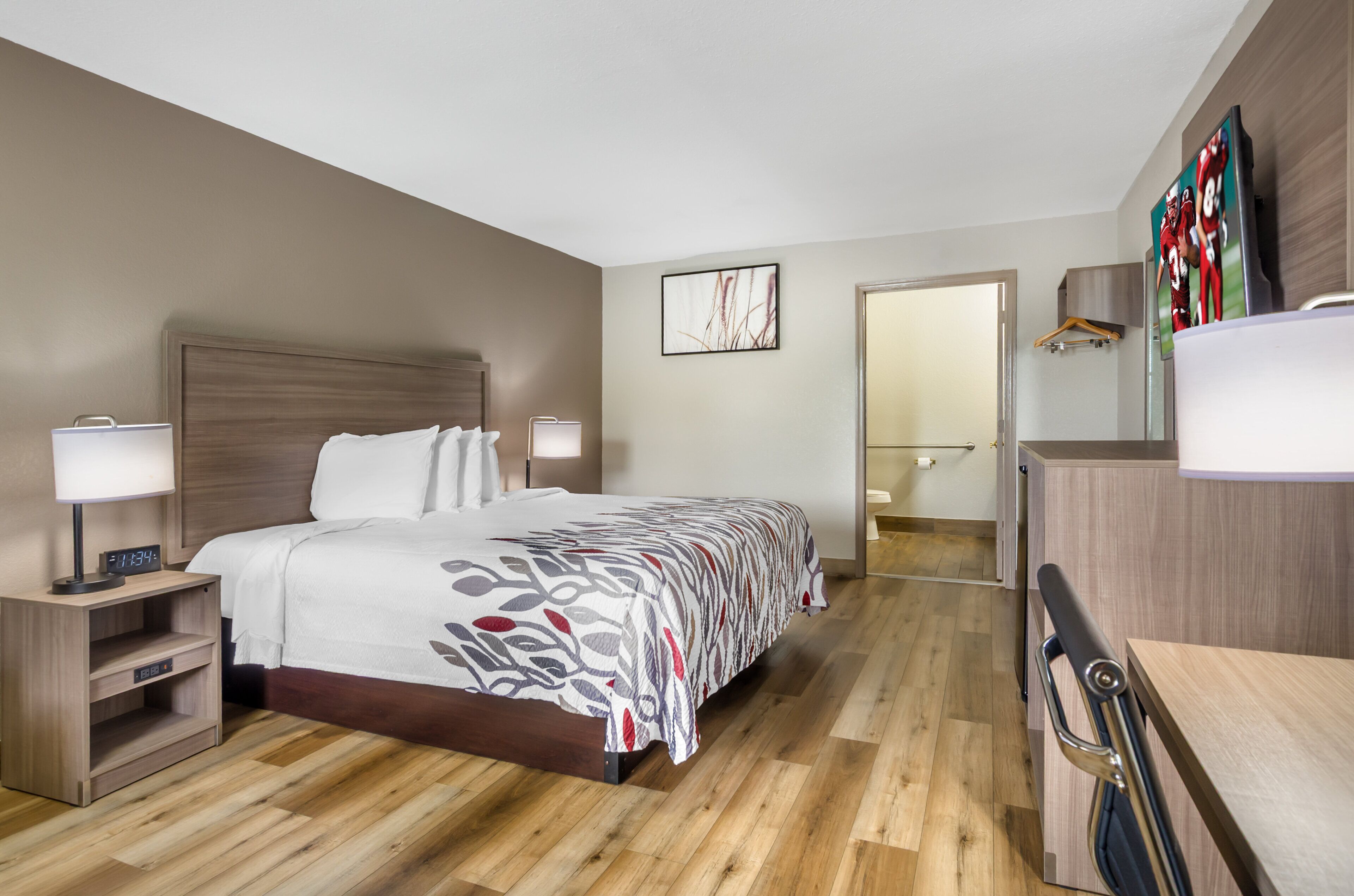 Een hotelkamer met een houten vloer, een bed met een beddeken met dessin, een nachtkastje met een lamp, een televisie aan de muur en een kapstok.