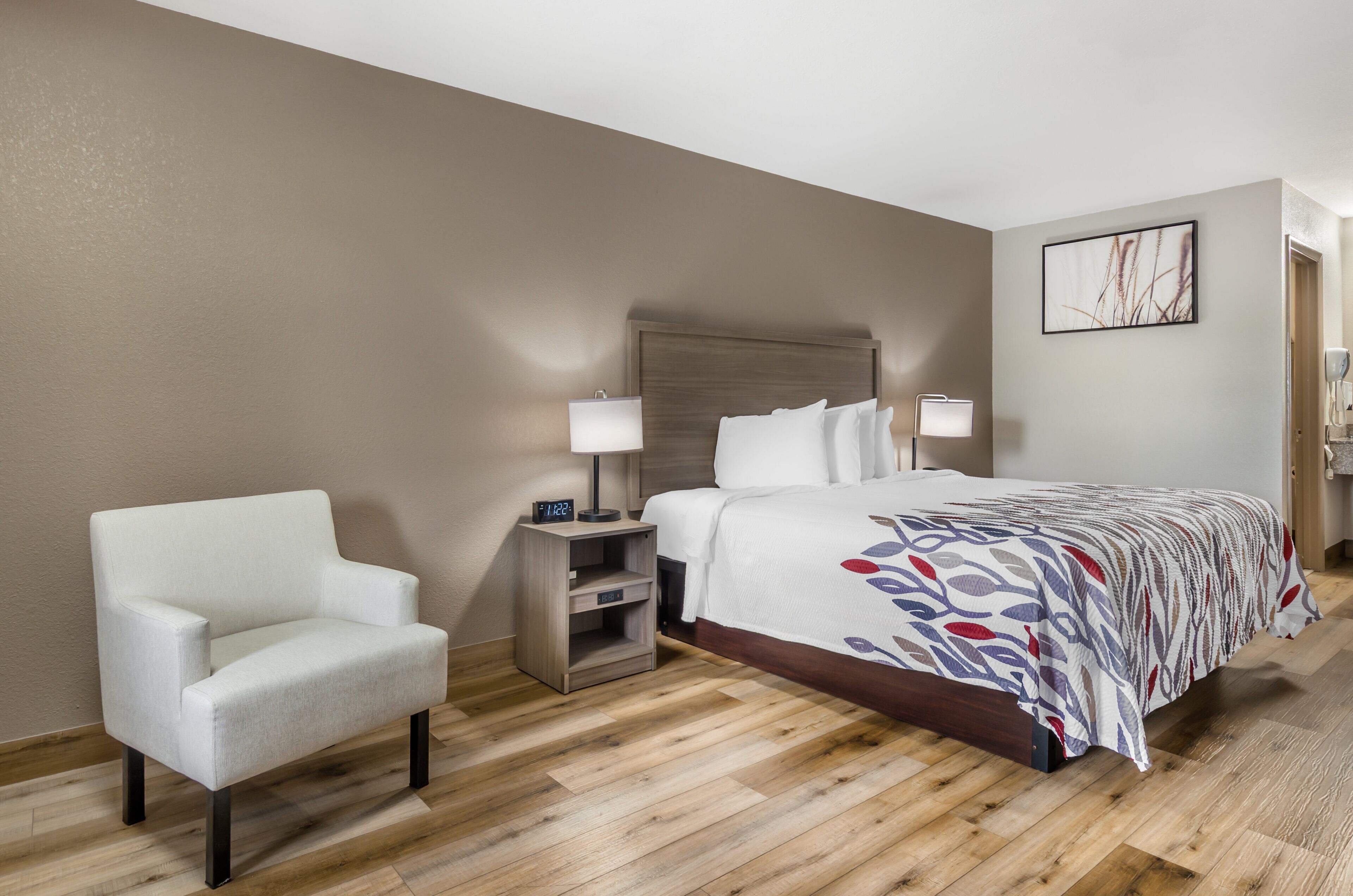 Una habitación de hotel con una cama, una silla, una mesita de noche con lámpara y un cuadro colgado en la pared.