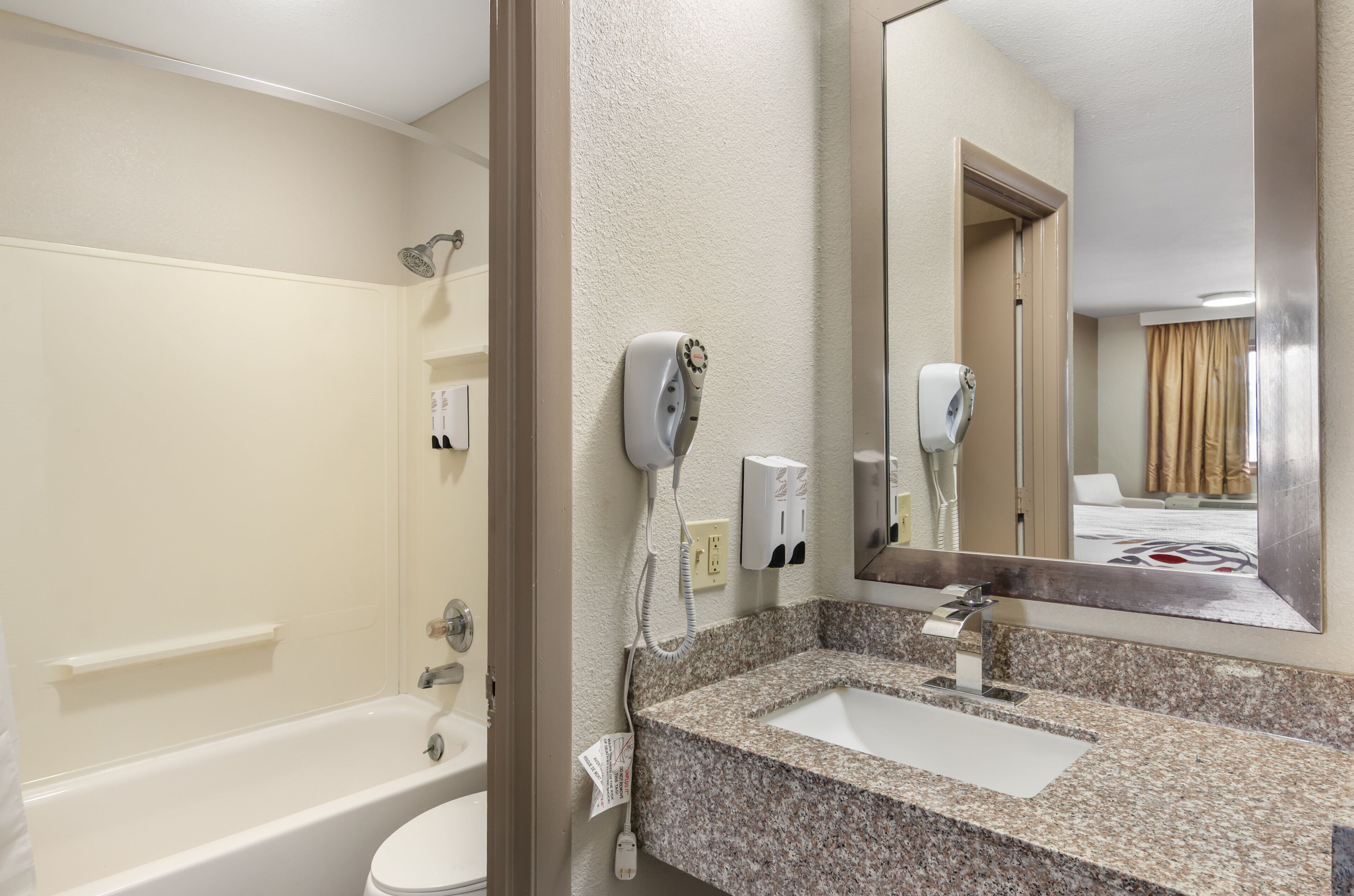 A hotel bathroom with a bathtub, a sink with a granite countertop, a mirror, and a hairdryer mounted on the wall.