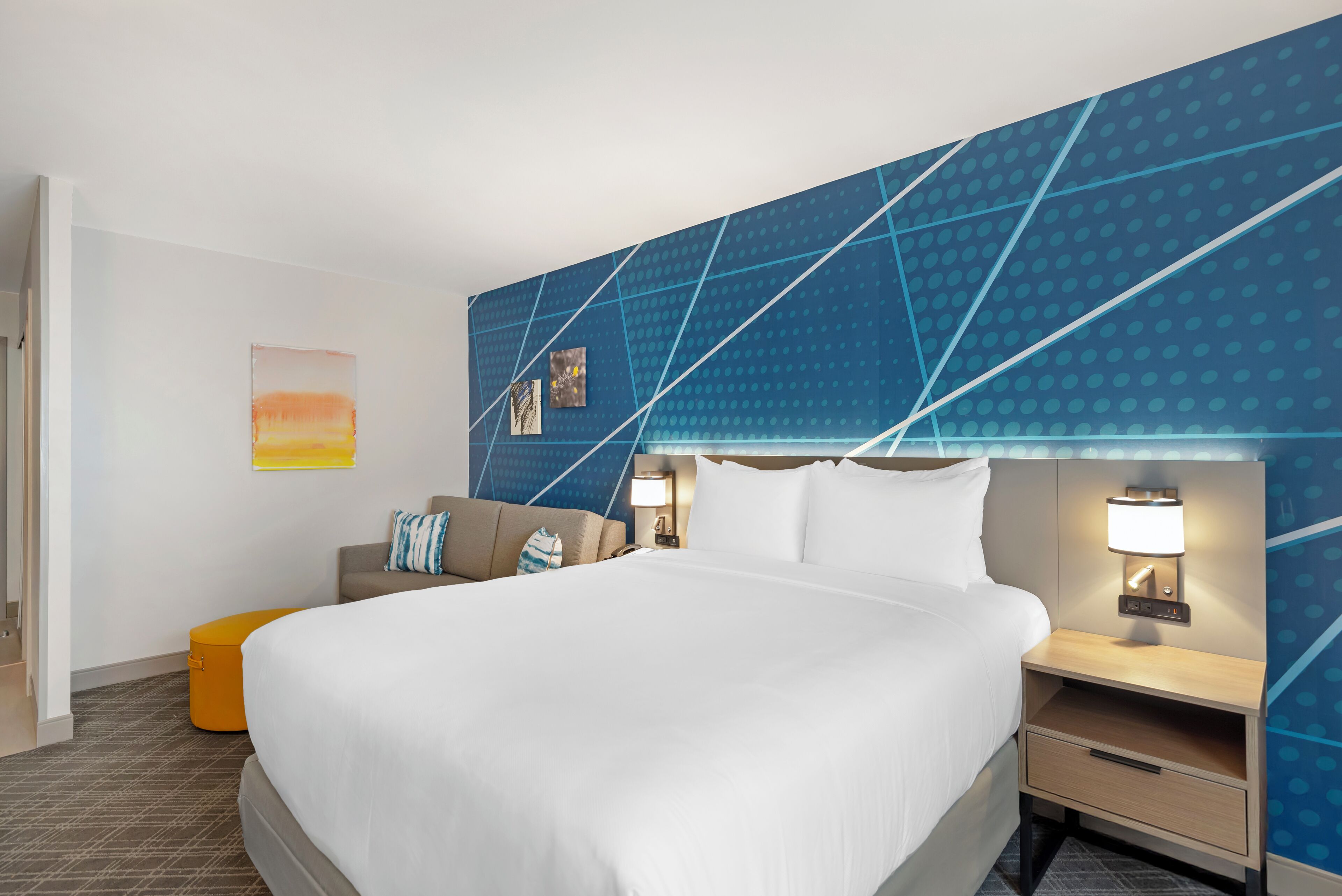 Ein Hotelzimmer mit einem großen Bett, einem Nachttisch mit Lampe, einem kleinen orangefarbenen Pouf und einer blauen Wand mit Muster.