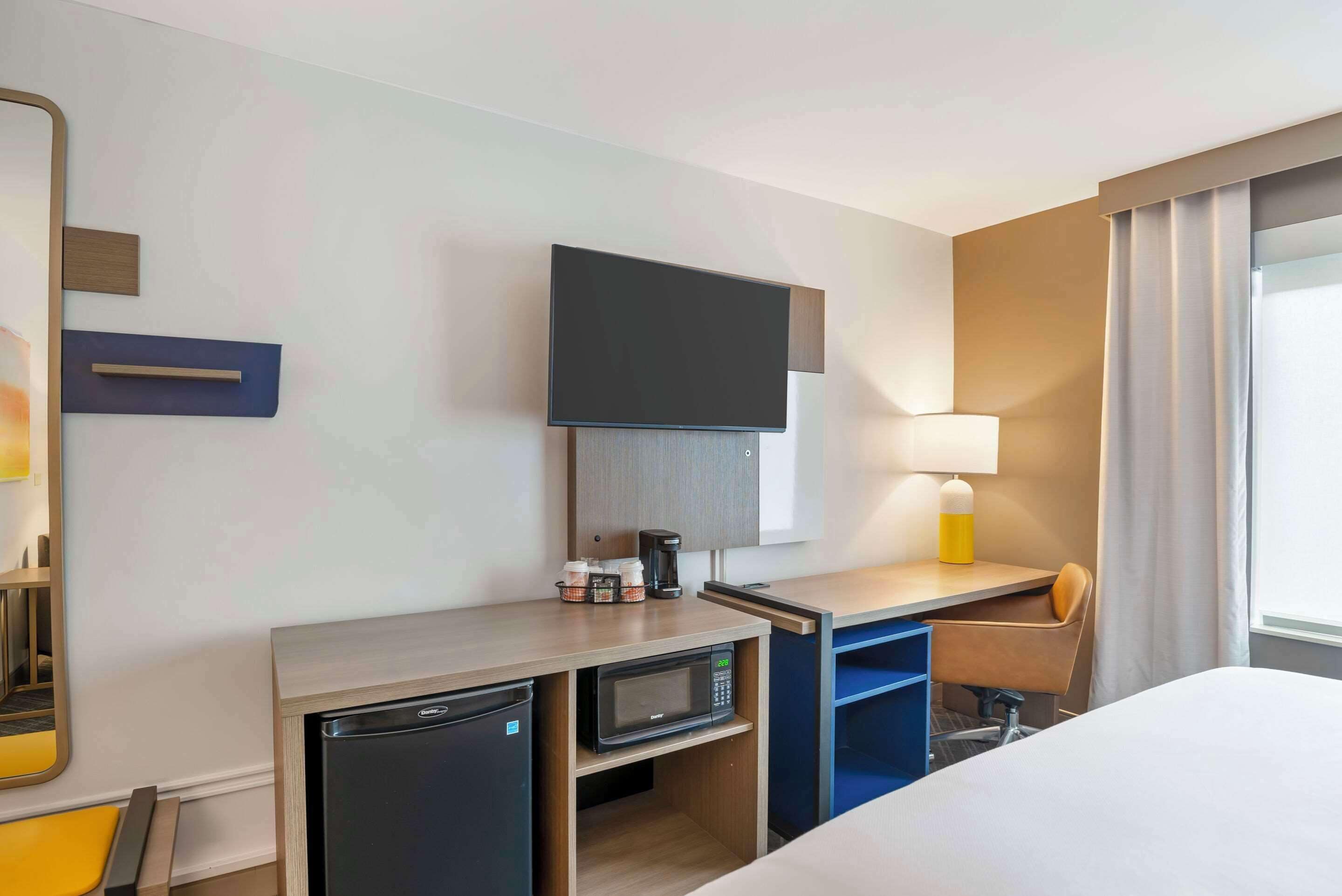 Une chambre d’hôtel équipée d’un lit, d’un bureau avec un micro-ondes, d’une télévision, d’une lampe et d’un petit réfrigérateur.