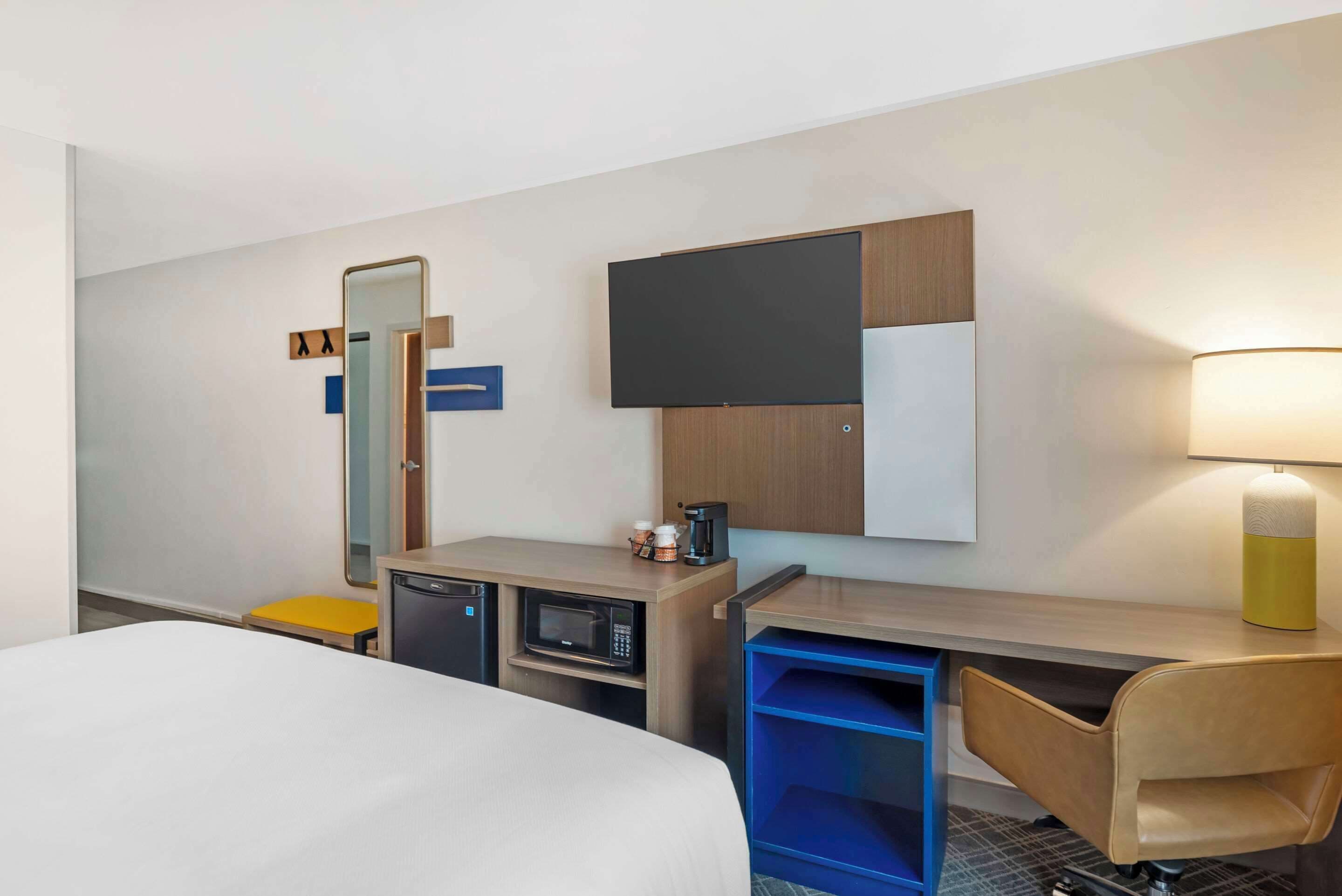 A hotel room with a bed, a desk with a computer, a microwave, a coffee maker, a lamp, and a mirror.