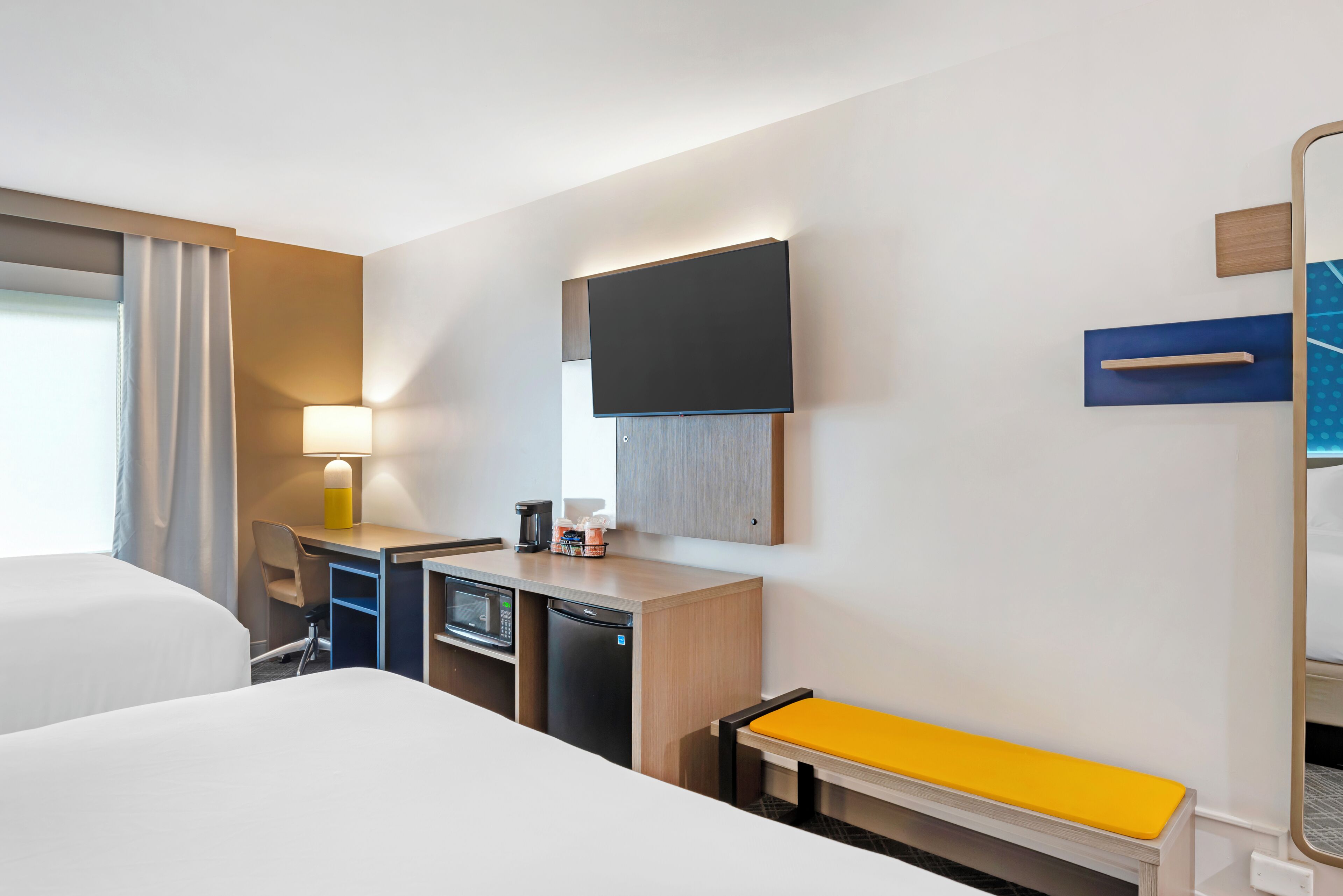 A hotel room with a bed, a desk with a computer, a microwave, a coffee maker, and a TV mounted on the wall.