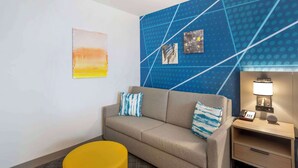 A hotel room with a beige sofa, two blue and white patterned pillows, a yellow ottoman, a blue accent wall with geometric patterns, and a small bedside table with a lamp and phone. - Comfort Inn Monticello (Charlottesville)