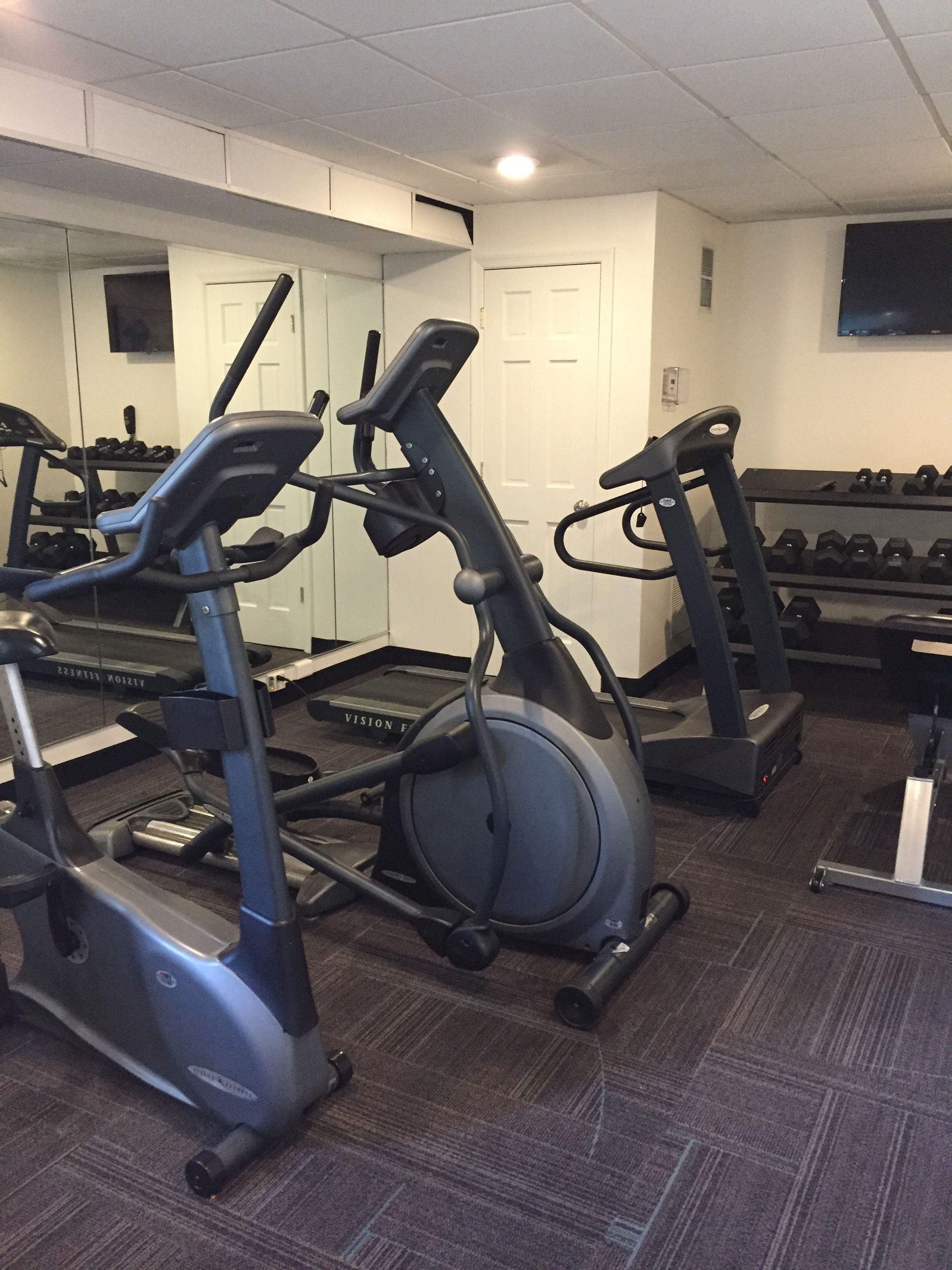 Une salle de sport équipée de plusieurs tapis de course, d’un porte-haltères et d’un appareil de musculation de la marque Vision.