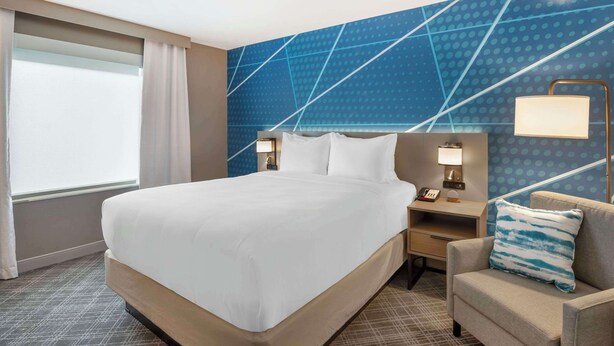 A hotel room with a large bed, a nightstand with a lamp, a chair, and a wall with a geometric pattern. - Comfort Inn Monticello (Charlottesville)