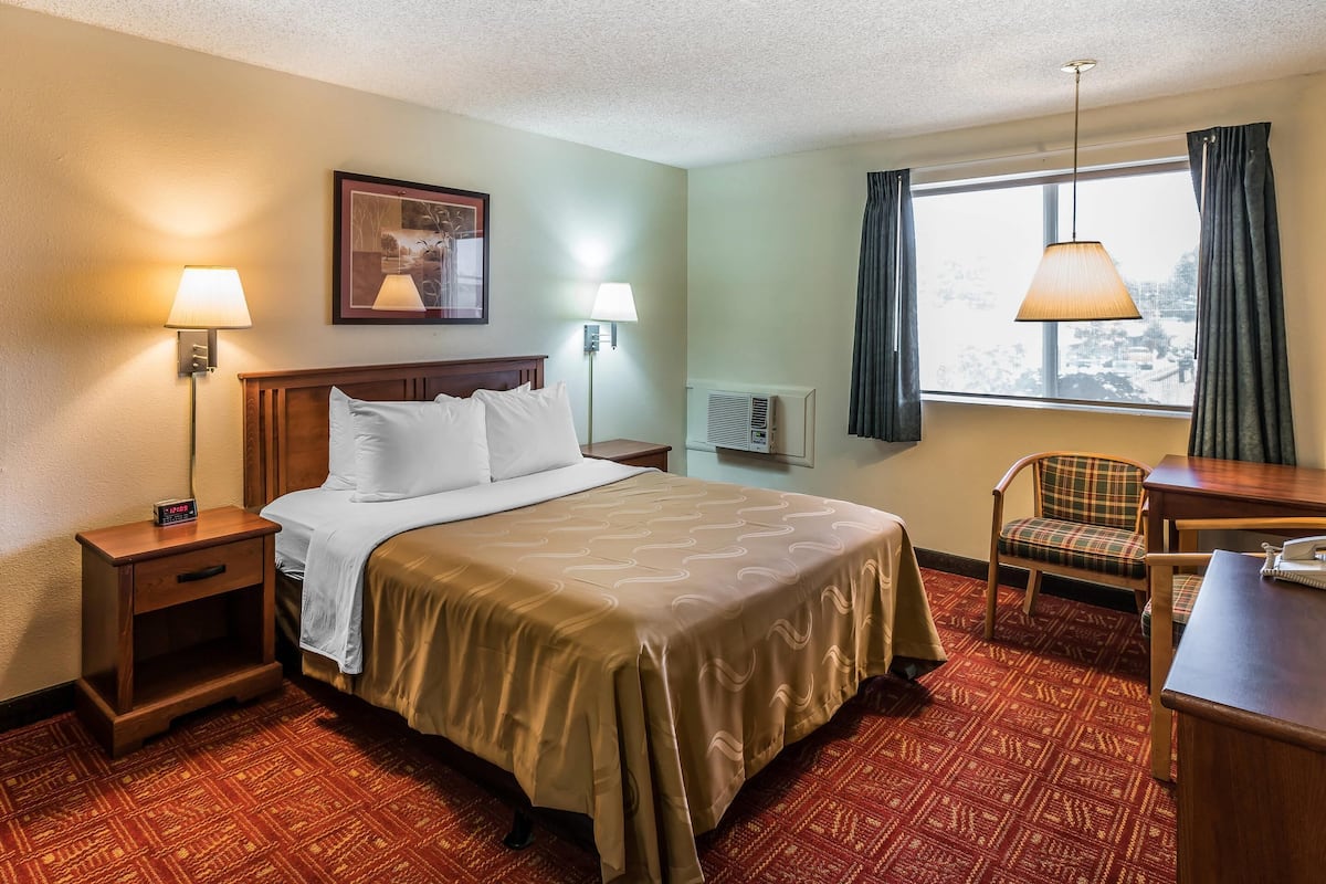 A hotel room with a bed, bedside lamps, a chair, a desk, and a window with curtains.