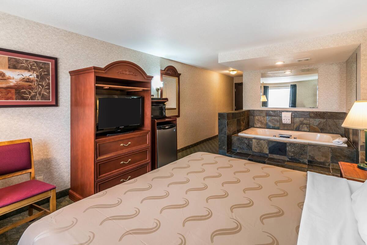 A hotel room with a bed, a TV, a desk, a chair, a bathroom with a bathtub, and a mirror.