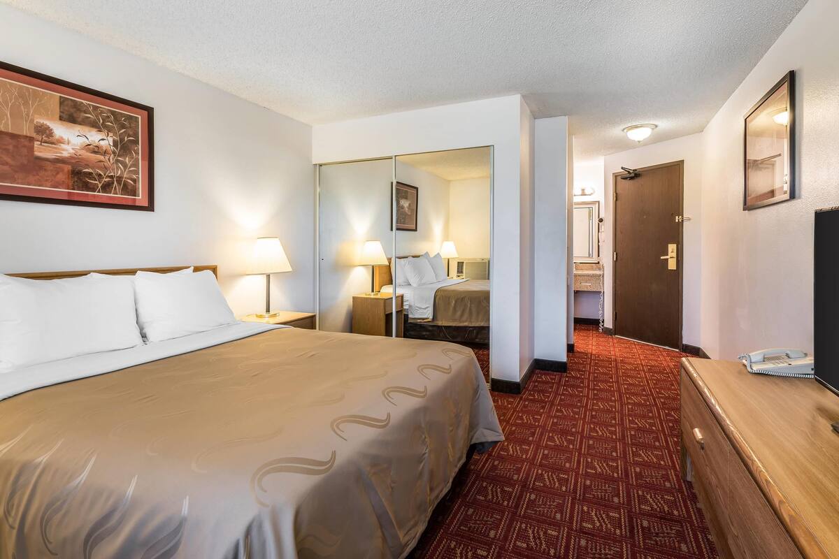 A hotel room with a large bed, a mirror, a wooden dresser, and a patterned carpet.