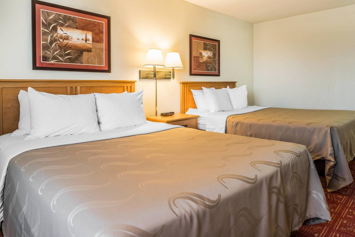 A hotel room with two beds, each with white linens and a patterned bedspread. There are two wall-mounted lamps on either side of the beds, and two framed artworks above them.