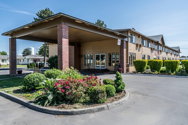 A motel with a covered entrance, a brick facade, and a landscaped front yard with various plants and shrubs.