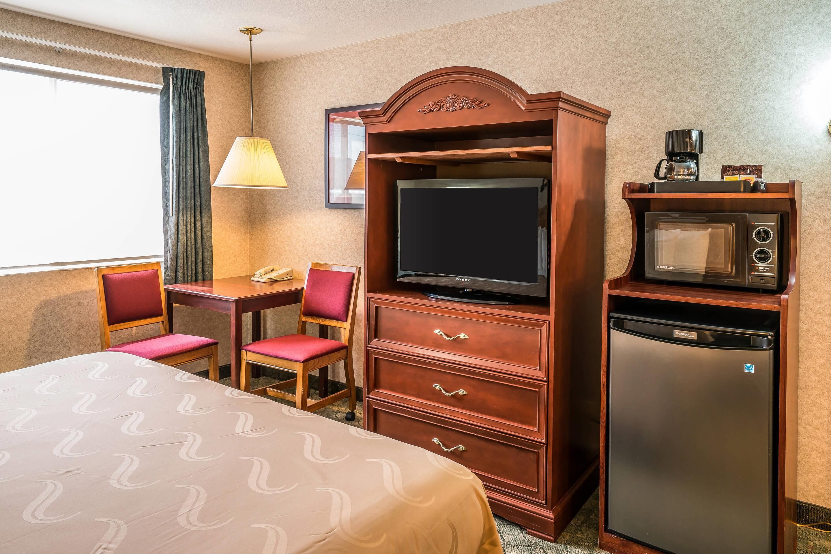 Ett hotellrum med en säng, ett skrivbord med en stol, en tv, en mikrovågsugn och ett kylskåp.