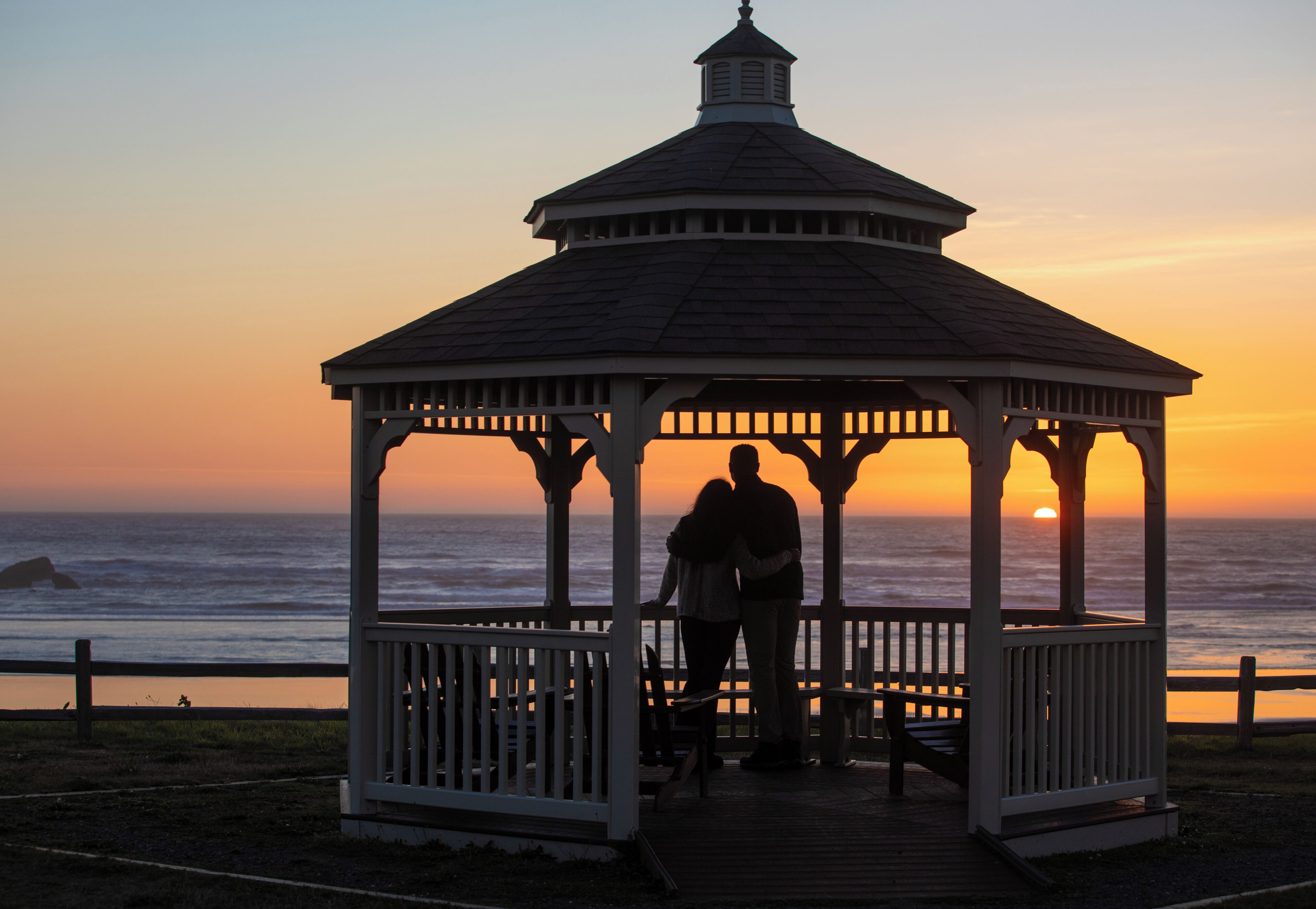 A gazebo with a white roof and wooden structure, overlooking the ocean at sunset.