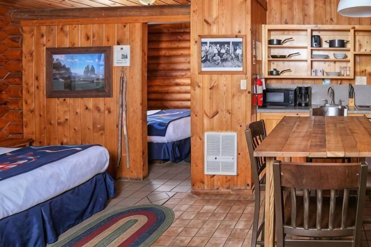 Une chambre située dans un chalet en rondins, avec deux lits, une table à manger en bois et un coin cuisine doté d’étagères.