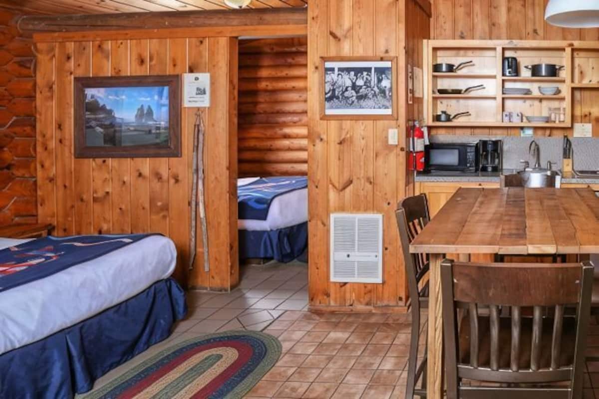 A log cabin room with two beds, a wooden dining table, and a kitchen area with shelves.