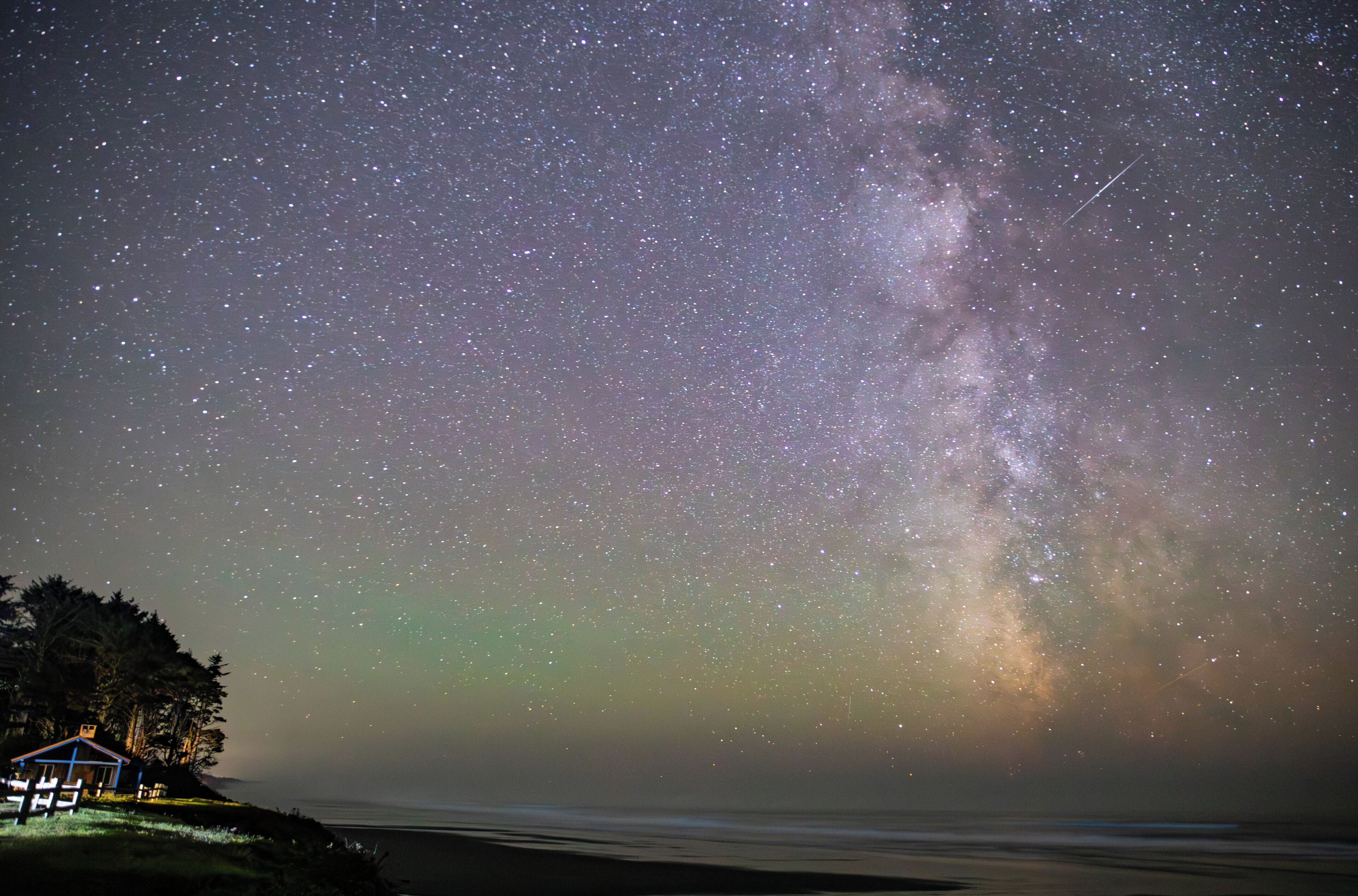 A starry night sky with a visible Milky Way, a beach, and a small illuminated structure.