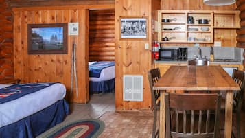 A cabin interior with a wooden dining table, two beds, a kitchenette, and a framed photo on the wall.