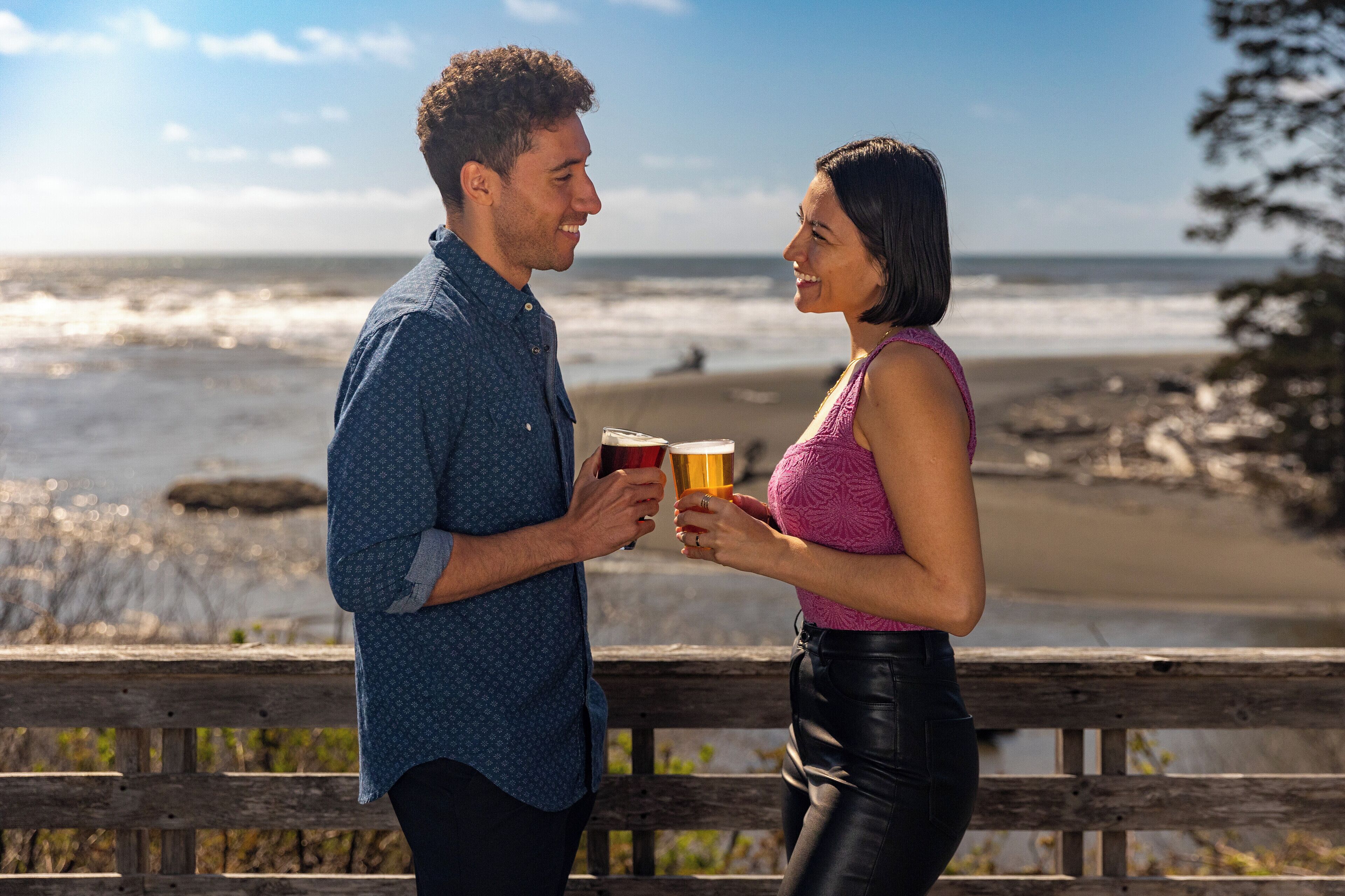 Two people holding drinks on a wooden deck with a beach and ocean in the background.