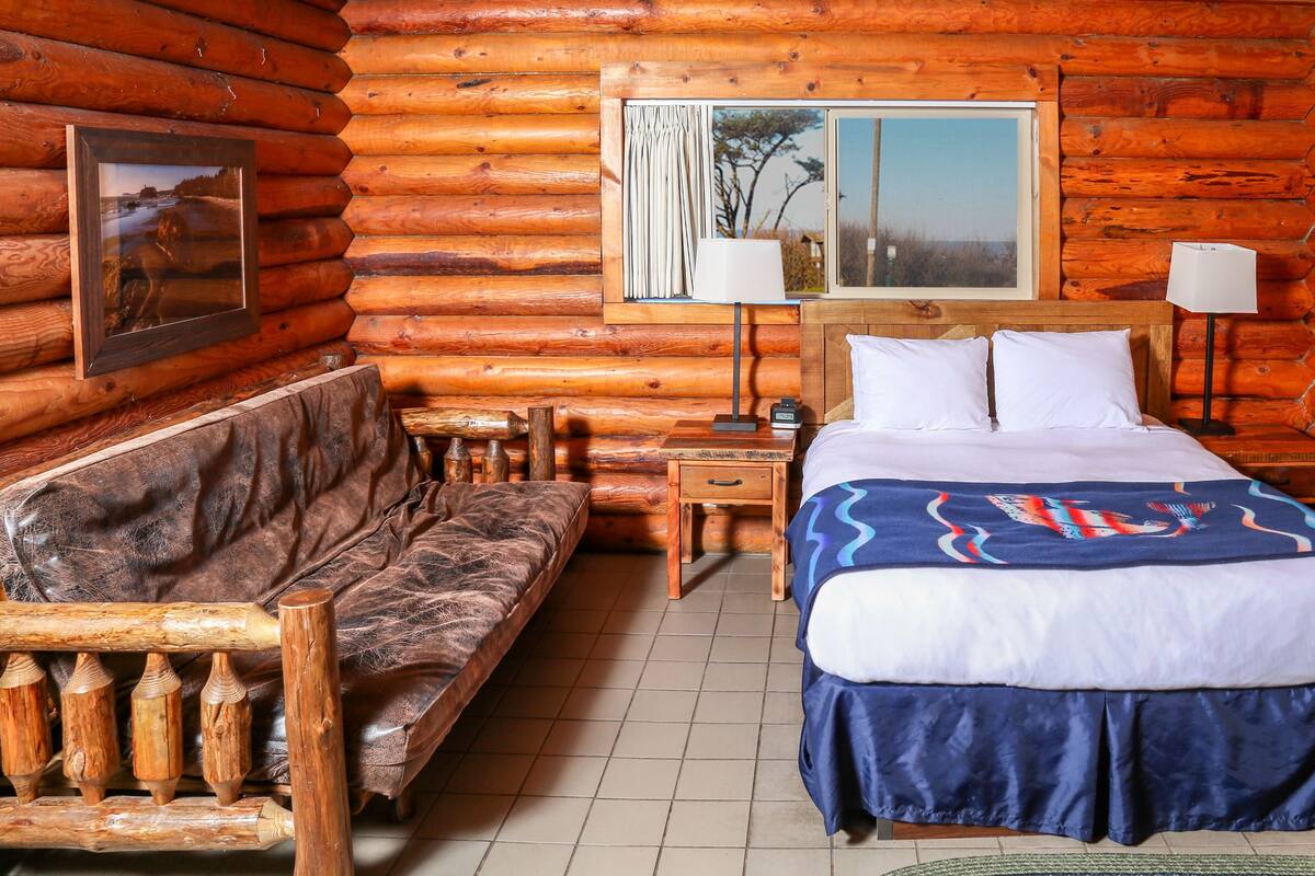 Une chambre aménagée dans un chalet en rondins, avec un lit, un banc en bois, une table de chevet, une lampe et un tableau encadré au mur.