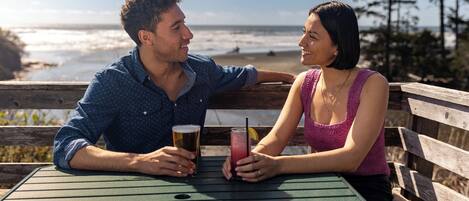 Dos personas sentadas a una mesa con bebidas, con vista a una playa y al océano.