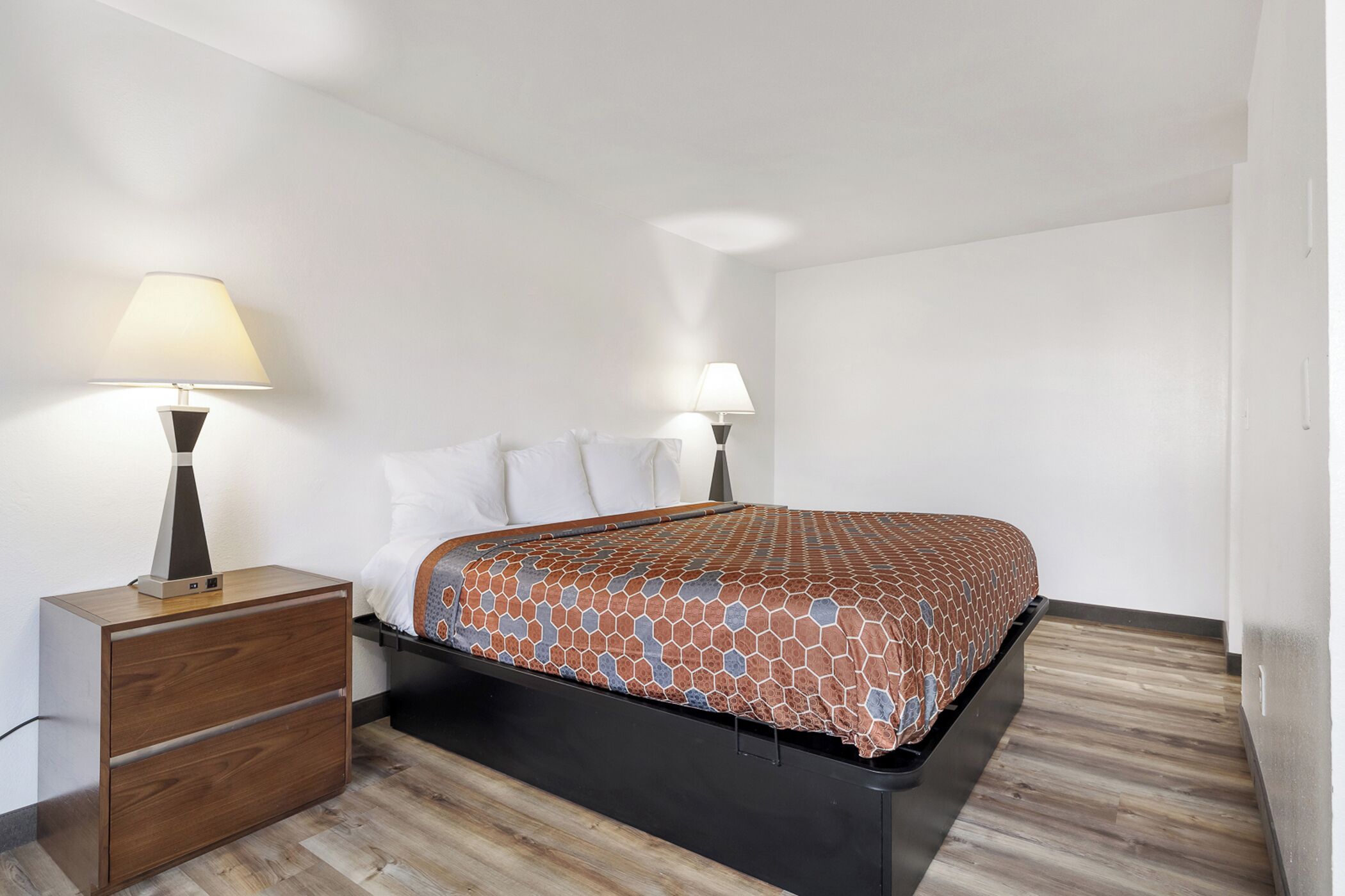 A bedroom with a bed, two bedside lamps, a wooden nightstand with drawers, and a patterned bedspread.