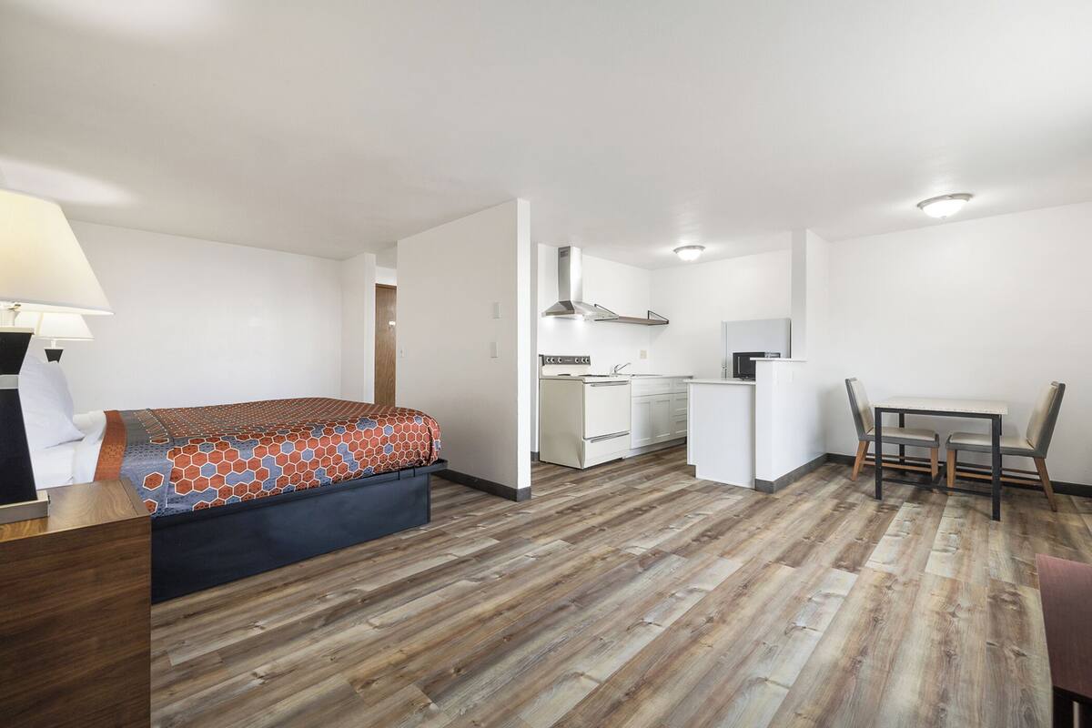 A compact hotel room with a bed, a desk, a chair, a small kitchenette, and a wooden floor.
