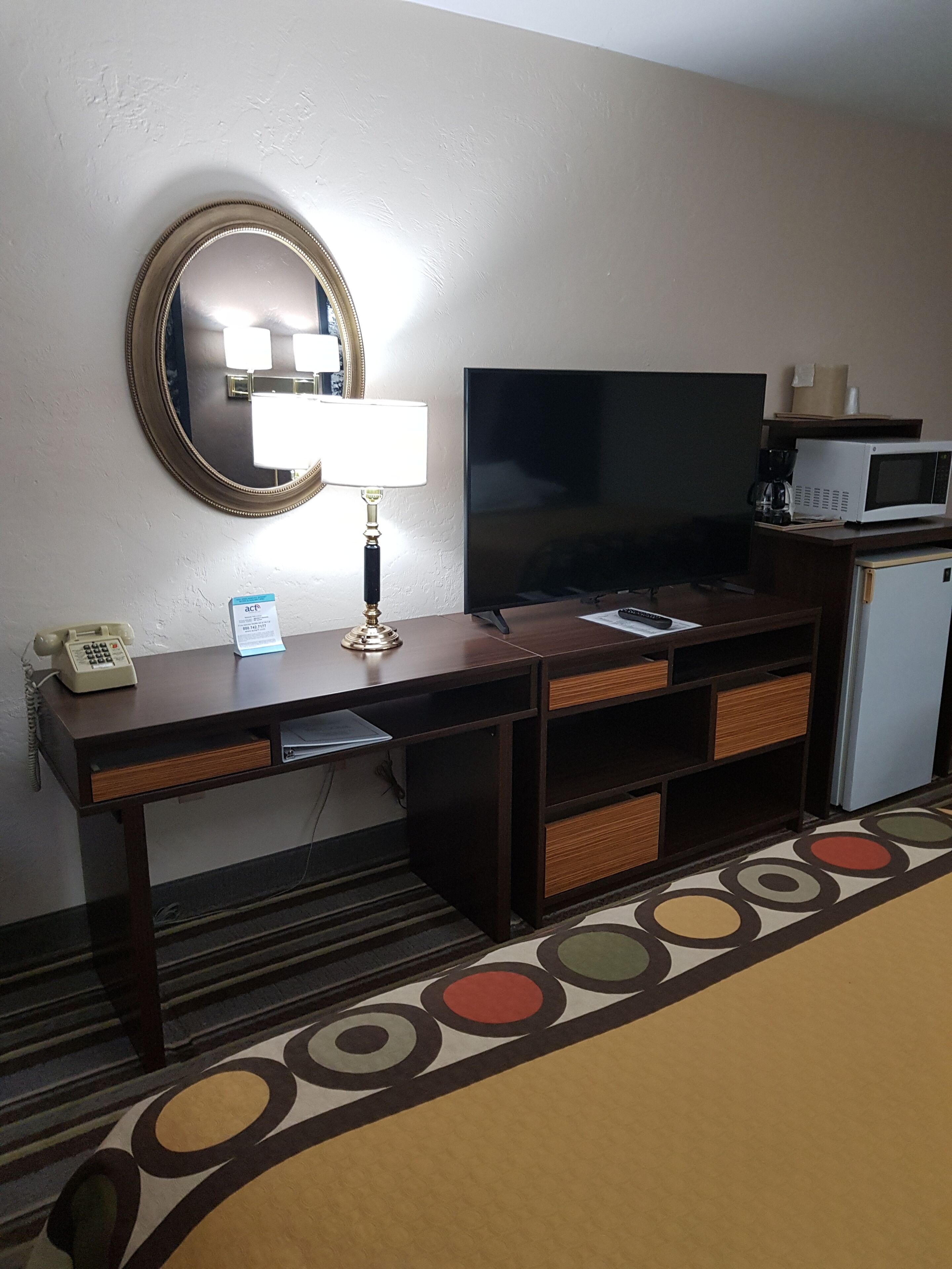 A hotel room with a wooden desk, a flat-screen TV, a microwave, a telephone, and a mirror on the wall.
