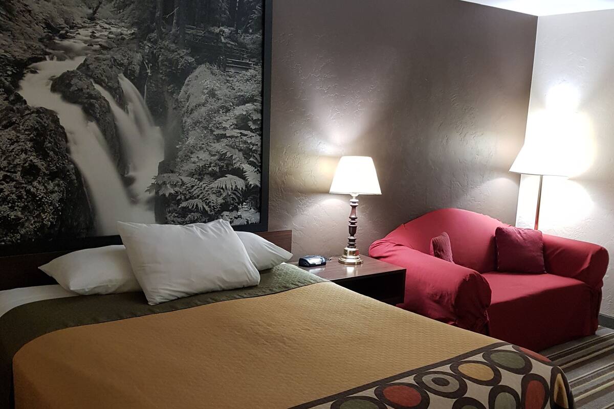 Ein Hotelzimmer mit einem großen Bett, einem roten Sessel, einem Nachttisch mit Lampe und einem Wandbild eines Wasserfalls.