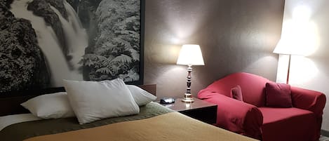 Una habitación de hotel con una cama grande, una silla roja, una mesita de noche con una lámpara y un cuadro colgado en la pared que representa una cascada.