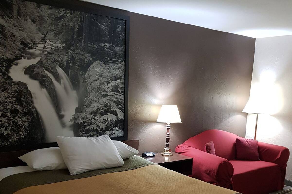Ein Hotelzimmer mit einem Bett, einem roten Sessel, einem Nachttisch mit Lampe und einem großen, gerahmten Bild einer Wasserfall.
