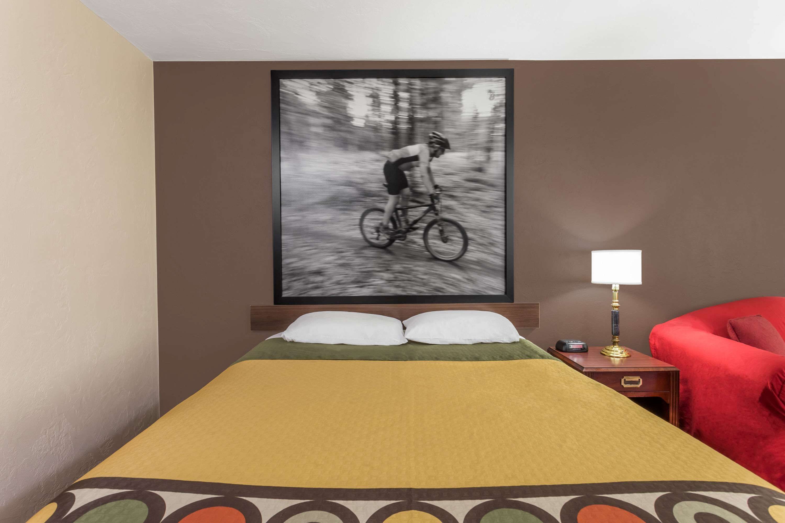 A hotel room with a bed, a framed picture of a cyclist, a nightstand with a lamp, and a red chair.