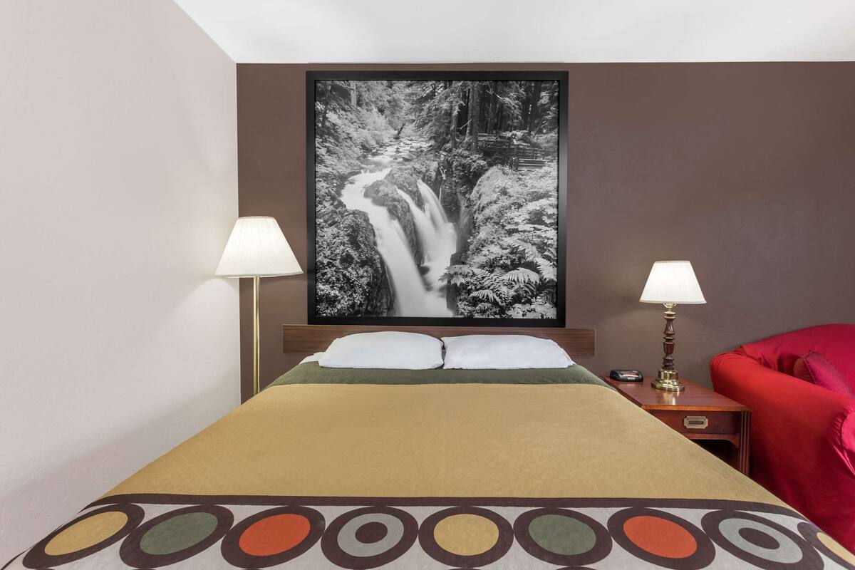 Ein Hotelzimmer mit einem Bett, zwei Nachttischlampen, einem roten Sessel und einem großen, gerahmten Bild einer Wasserfälle an der Wand.