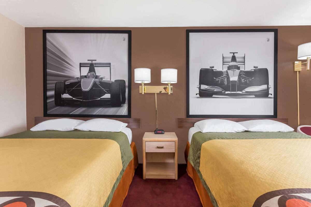Ein Hotelzimmer mit zwei Betten, jedes mit einer grünen und gelben Tagesdecke, einem Nachttisch mit Lampe sowie zwei gerahmten Bildern von Rennwagen an der Wand.