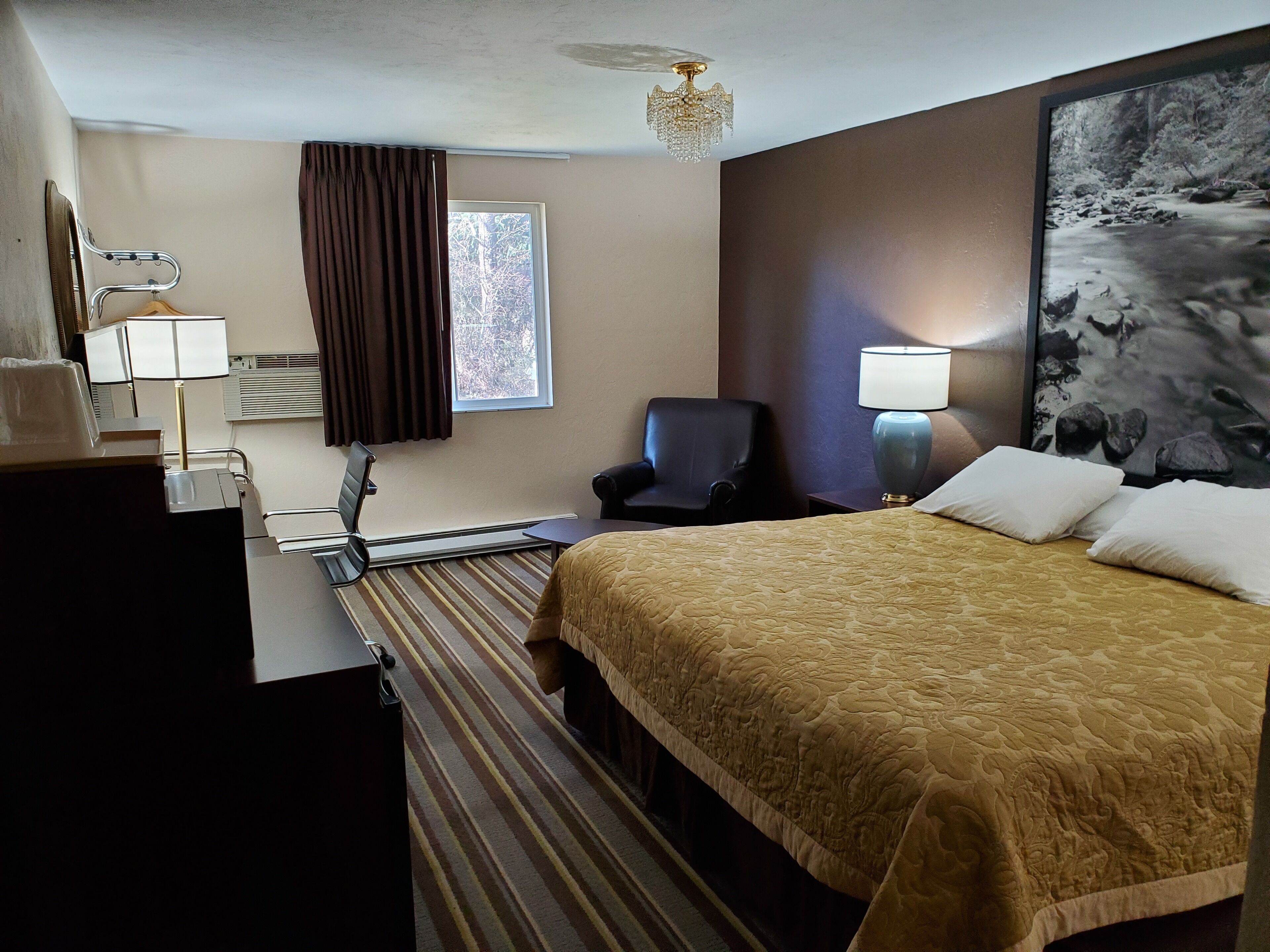 Una habitación de hotel con cama, escritorio con lámpara, silla, ventana con cortinas y un cuadro en la pared.