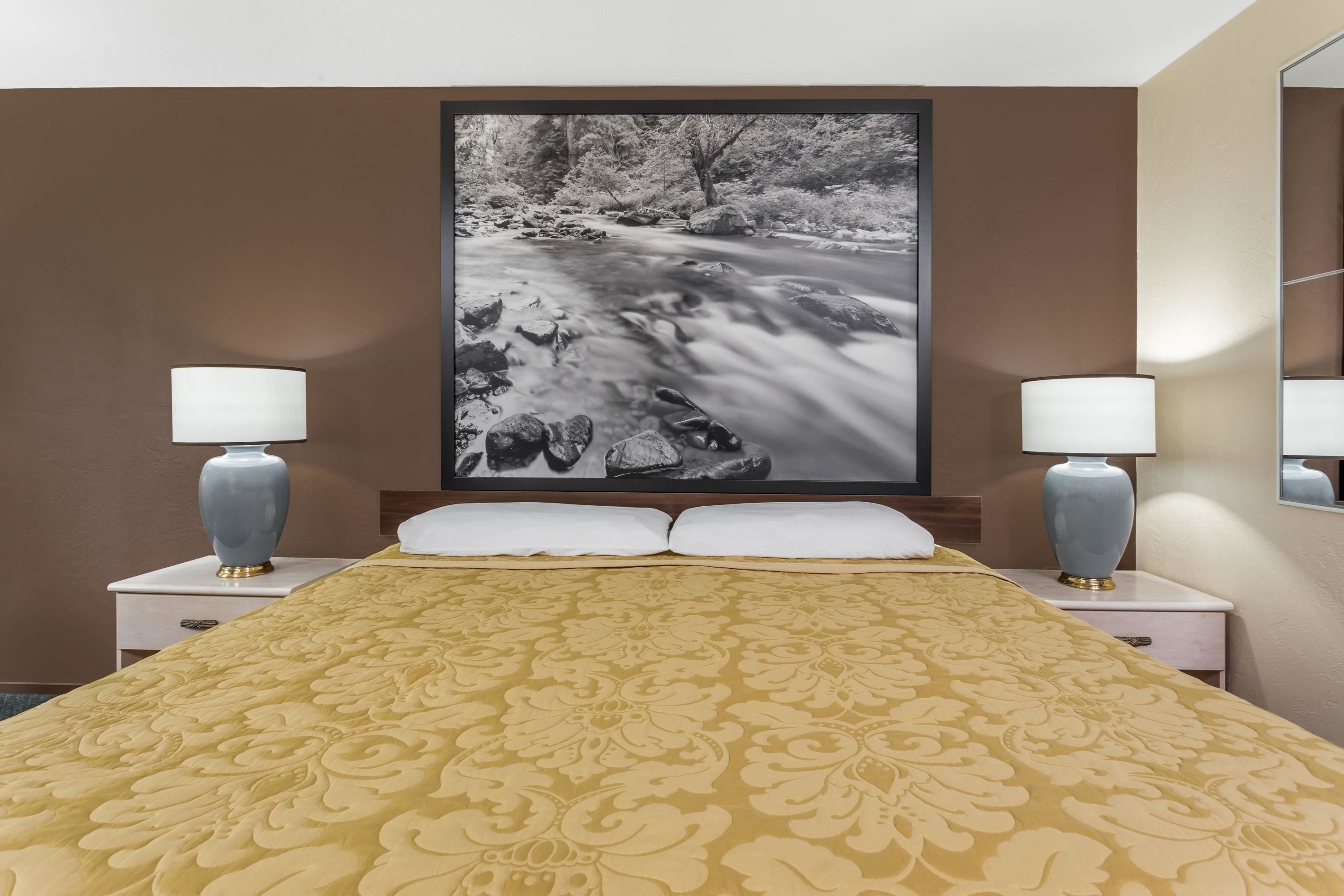 Une chambre d’hôtel avec un grand lit, deux tables de chevet avec des lampes et un tableau représentant un paysage fluvial accroché au mur.