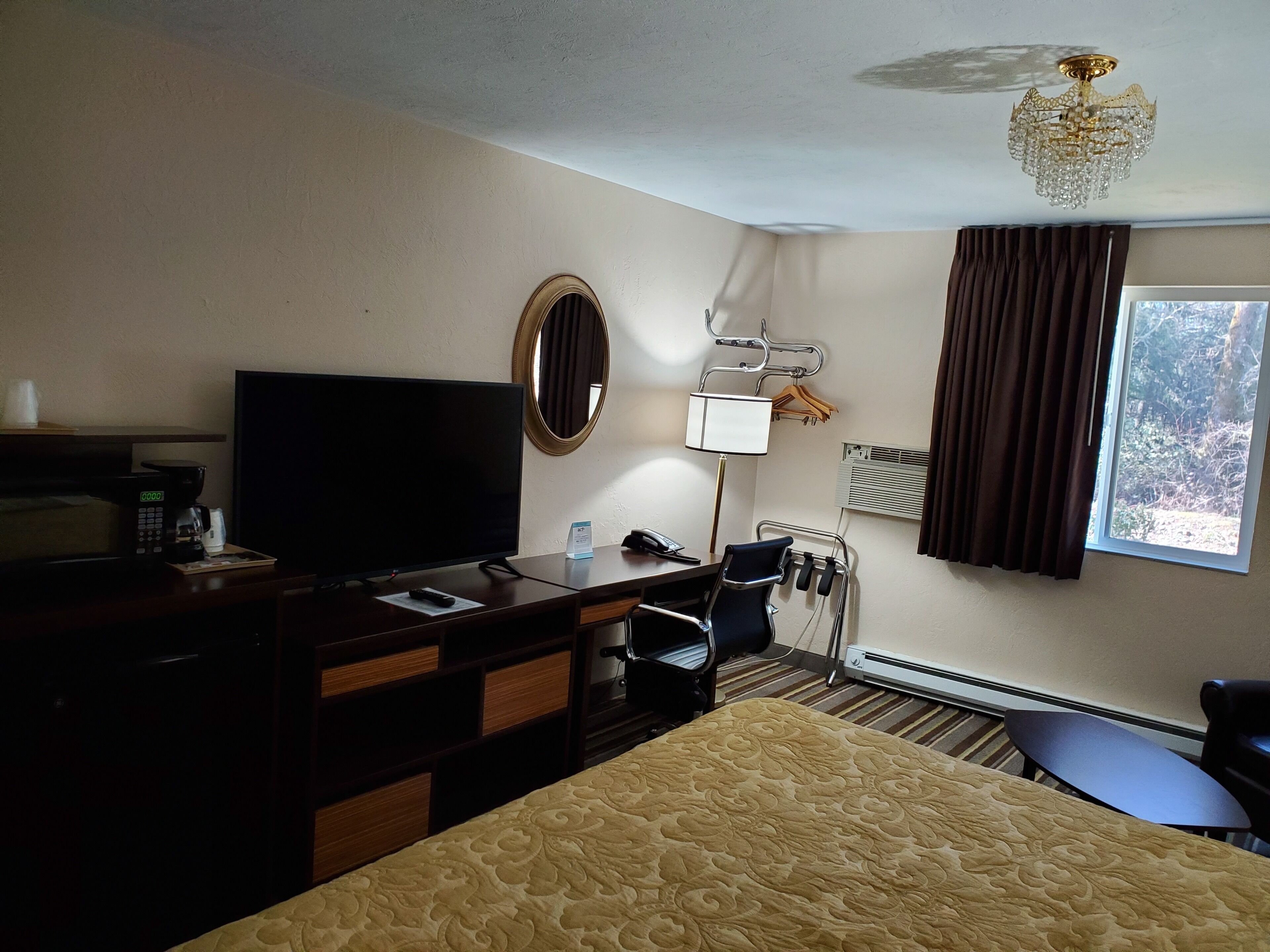 A hotel room with a bed, a desk with a coffee maker, a TV, a mirror, a lamp, a heater, and a window with curtains.